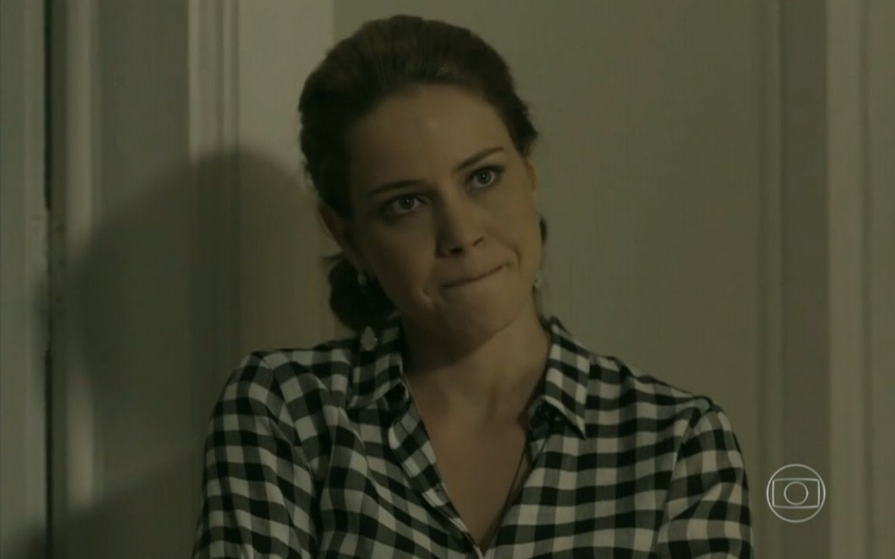 Cristina (Leandra Leal) está em pé próximo à porta de sua sala em cena de Império; ela usa uma blusa xadrez preta e branca