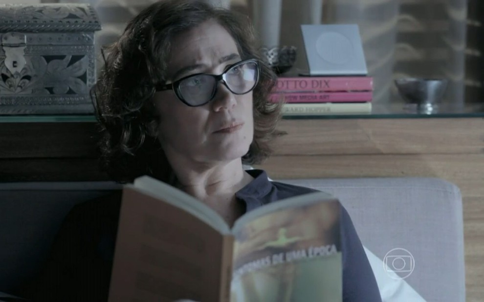 Lilia Cabral grava com livro na mão, óculos de grau, deitada na cama e olhando para o lado com expressão séria como Maria Marta