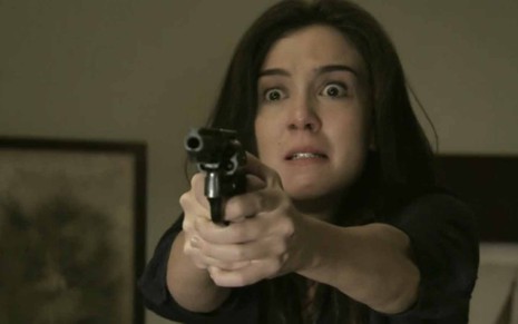 A atriz Marjorie Estiano como Cora segura um revólver com as duas mãos e arregala os olhos em cena de Império