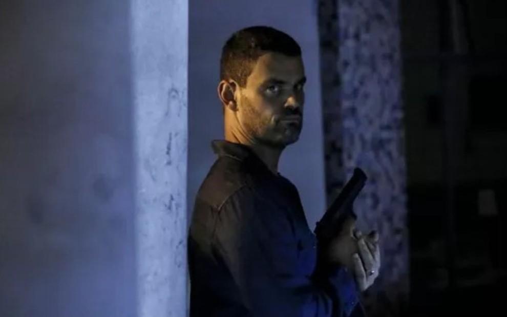 O ator Carmo Dalla Vecchia com expressão séria, segurando uma arma em cena de Império em cenografia escura, à noite