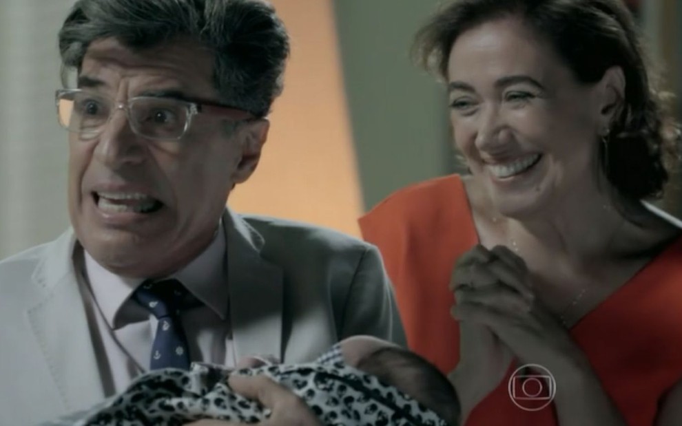 Téo Pereira (Paulo Betti) segura bebê no colo e  Maria Marta (Lilia Cabral) sorri atrás dele em cena de Império