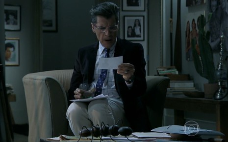 Paulo Betti grava de terno e óculos de grau sentado em uma poltrona, onde lê um papel como Téo de Império