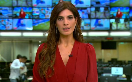 Andréia Sadi usa uma camisa vermelha e encara a câmera, séria, no estúdio do Jornal Hoje