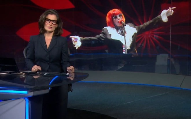 Renata Vasconcellos está na bancada do Jornal Nacional, com uma imagem de Rita Lee ao fundo