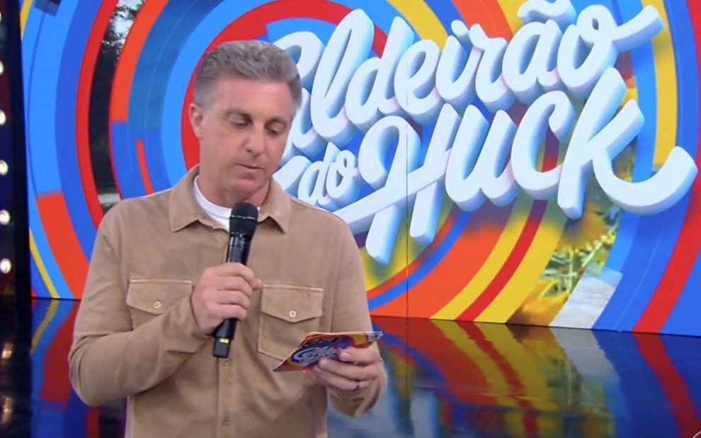 Luciano Huck, vestido com uma camiseta marrom, no estúdio de fundo azul de seu programa na Globo que vai ao ar nas tardes de sábado