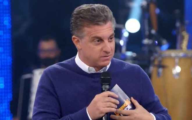 O apresentador Luciano Huck segura microfone e olha para frente com cara de sério no Domingão com Huck