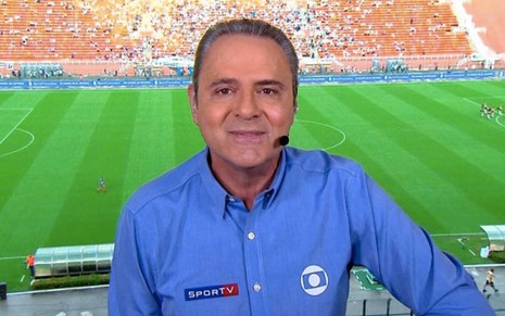 O narrador Luis Roberto com uma camisa azul, de manga longa, em frente a um gramado verde de um estádio