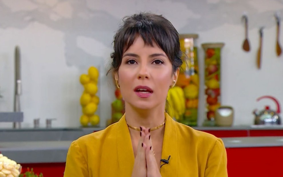 Andréia Horta está com as mãos coladas uma na outra, usa roupa amarela e está sentada na mesa do café da manhã do Mais Você