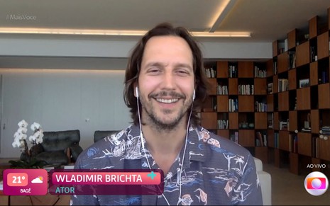 Vladimir Brichta usa camisa florida e sorri para a câmera; na frente, é possível ver o GC, no qual está inscrito "Wladimir Brichta".