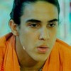 Vestindo colete laranja neon e com cabelos compridos amarrados, Andre Marques, caracterizado como Mocotó, olha para além da câmera em cena de Malhação