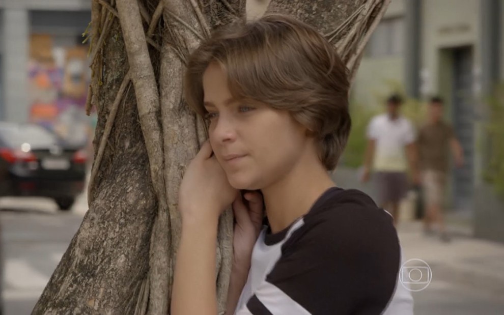 Isabella Santoni grava abraçada em uma árvore com camiseta branca e preta e expressão triste como Karina