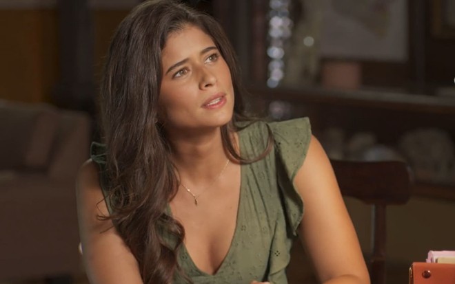 Isadora Cruz, caracterizada como Candoca, tem a expressão séria e pensativa; atriz tem os cabelos longos presos em um rabo de cavalo baixo e veste uma camisa laranja