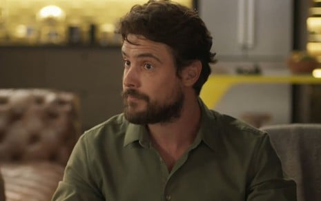 Sergio Guizé usa uma camisa verde e tem os cabelos bem arranjados e a barba arrumada; ele ostenta uma expressão confusa, com os olhos arregalados, em cena de Mar do Sertão