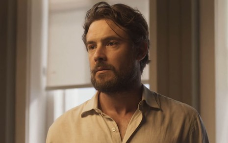 Sergio Guizé caracterizado como José; ator usa uma camisa branca e uma corrente de prata e dá um sorriso irônico em cena de Mar do Sertão