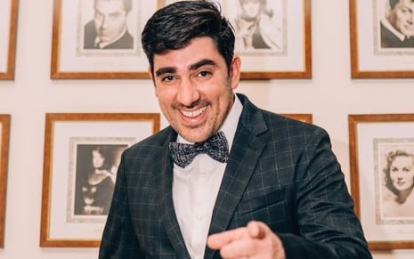 Marcelo Adnet sorri e aponta para a câmera; ele usa um smoking azulado com estampa xadrez e gravata borboleta