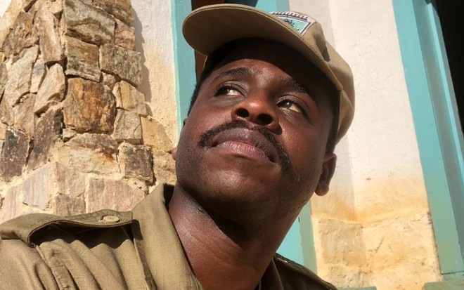 Hugo Germano usa o uniforme do soldado Venâncio --blusa e boné beges-- e encara um ponto fixo; a foto foi tirada de baixo para cima, e ele está sério