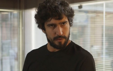 Renato Góes, caracterizado como Tertulinho, encara um ponto fixo com deboche; ator tem os olhos semicerrados, a boca em um sorriso irônico e veste uma camisa preta