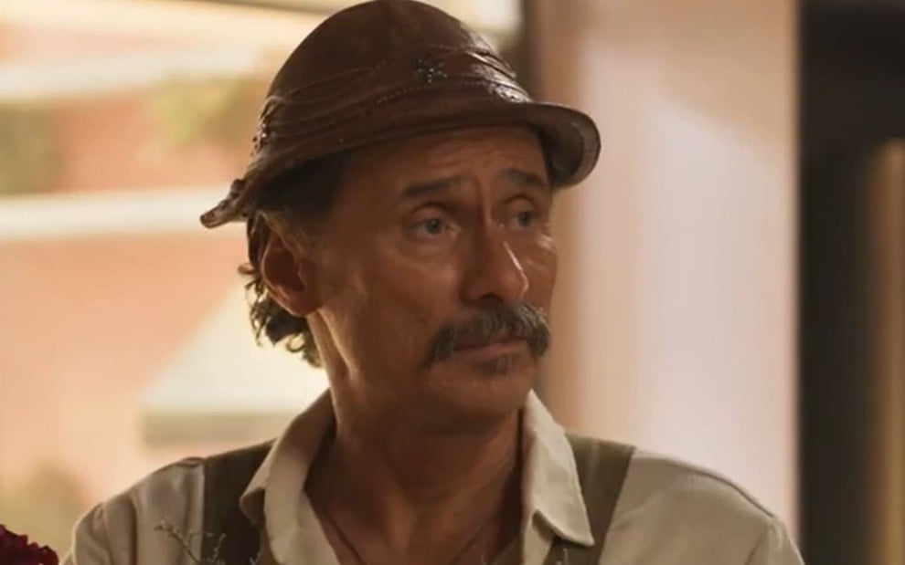 Enrique Diaz caracterizado como Timbó; ele veste roupas beges e surradas e exibe choque em cena de Mar do Sertão