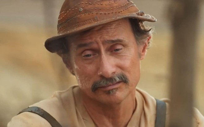 Enrique Diaz usa roupas beges e gastas e um gibão; caracterizado como Timbó, ator dá um sorriso cínico em cena de Mar do Sertão