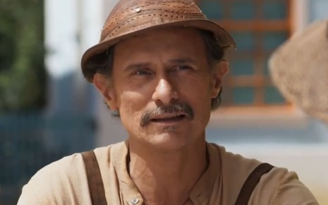 Enrique Diaz caracterizado como Timbó, ele veste roupas beges e surradas e tem a boca levemente aberta em cena de Mar do Sertão