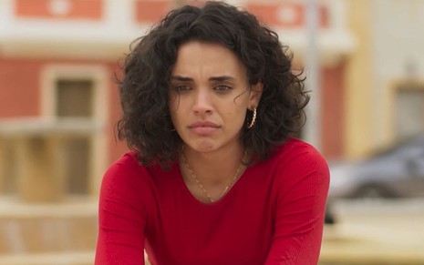 Giovana Cordeiro usa os cabelos cacheados na altura dos ombros e veste um conjunto vermelho; ela está desolada, com os olhos cheios de lágrimas, em cena de Mar do Sertão