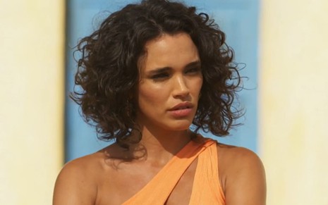Giovana Cordeiro caracterizada como Xaviera; atriz usa um vestido laranja e tem a expressão debochada em cena de Mar do Sertão