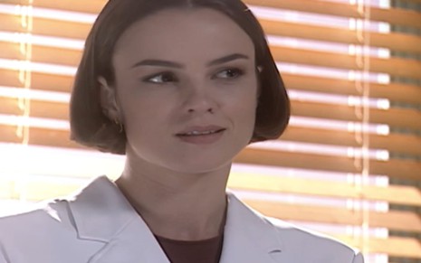 Carolina Kasting caracterizada como Laura; ela usa um jaleco branco e os cabelos curtos, na altura do queixo. A atriz dá um sorriso cúmplice em cena de Mulheres Apaixonadas