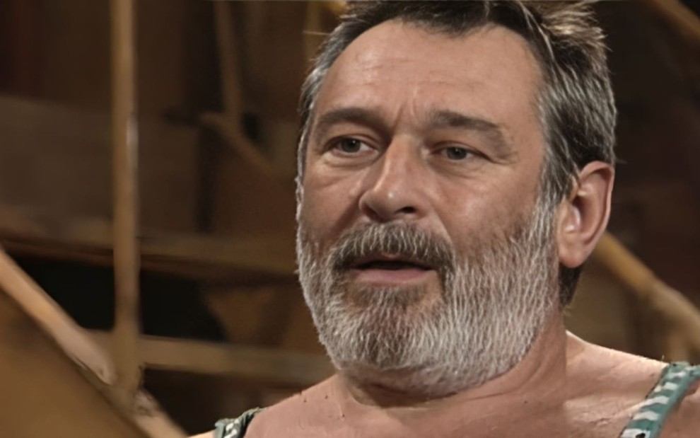 Paulo Goulart caracterizado como Donato; ele usa uma regata branca e uma boina marrom e dá um sorriso sacana em cena de Mulheres de Areia.
