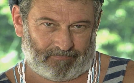 Paulo Goulart caracterizado como Donato; ele usa uma regata branca e uma boina marrom. Ele dá um sorriso sacana em cena de Mulheres de Areia.
