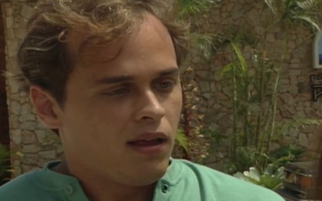 Guilherme Fontes caracterizado como Marcos; ele tem os cabelos castanhos curtos e usa uma camisa verde. O semblante exprime confusão em cena de Mulheres de Areia