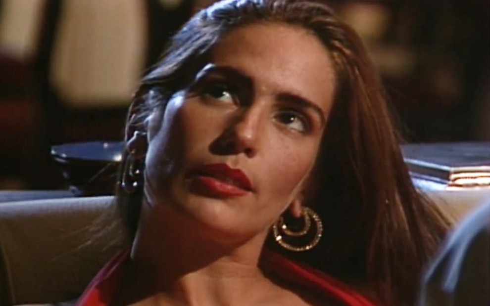 Gloria Pires caracterizada como Raquel; ela tem os cabelos soltos e longos e usa um vestido vermelho. O semblante está sério e perturbado em cena de Mulheres de Areia.
