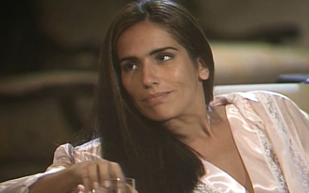 Gloria Pires caracterizada como Raquel; ela tem os cabelos soltos e longos e usa um vestido florido. O semblante está sério e perturbado em cena de Mulheres de Areia.