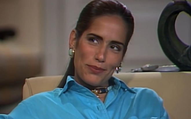 Gloria Pires caracterizada como Raquel; ela tem os cabelos soltos e longos e usa um vestido florido. O semblante está sério e perturbado em cena de Mulheres de Areia.