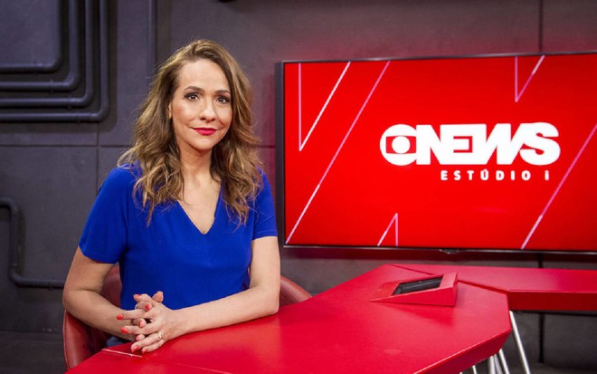 Maria Beltrão com uma blusa azul, de braços cruzados e sorrindo levemente para a câmera da Globo News, no estúdio do Estúdio I