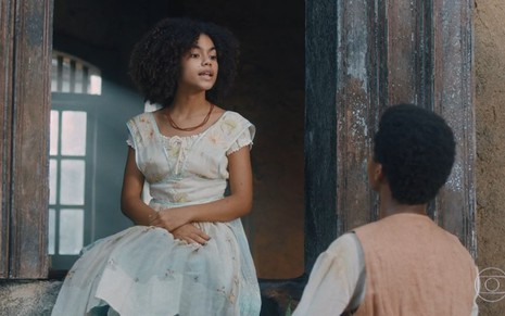 Alana Cabral grava com vestido branco, sentada na janela e conversando com um rapaz como Zayla na primeira fase de Nos Tempos do Imperador
