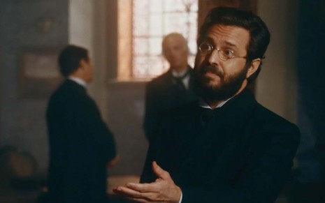 O ator Alcemar Vieira com óculos redondos, de aro metálico, e barba grande, preta, como José de Alencar em cena de Nos Tempos do imperador