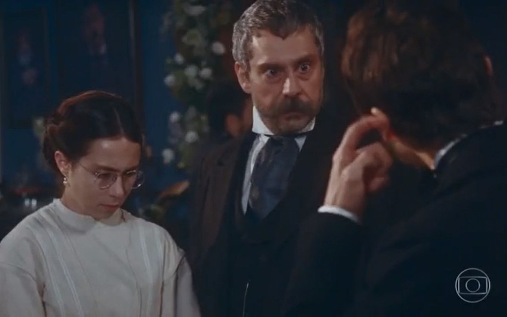Tonico (Alexandre Nero) está ao lado de Dolores (Daphne Bozaski) e conversa com Nélio (João Pedro Zappa), que está de costas na foto, em evento