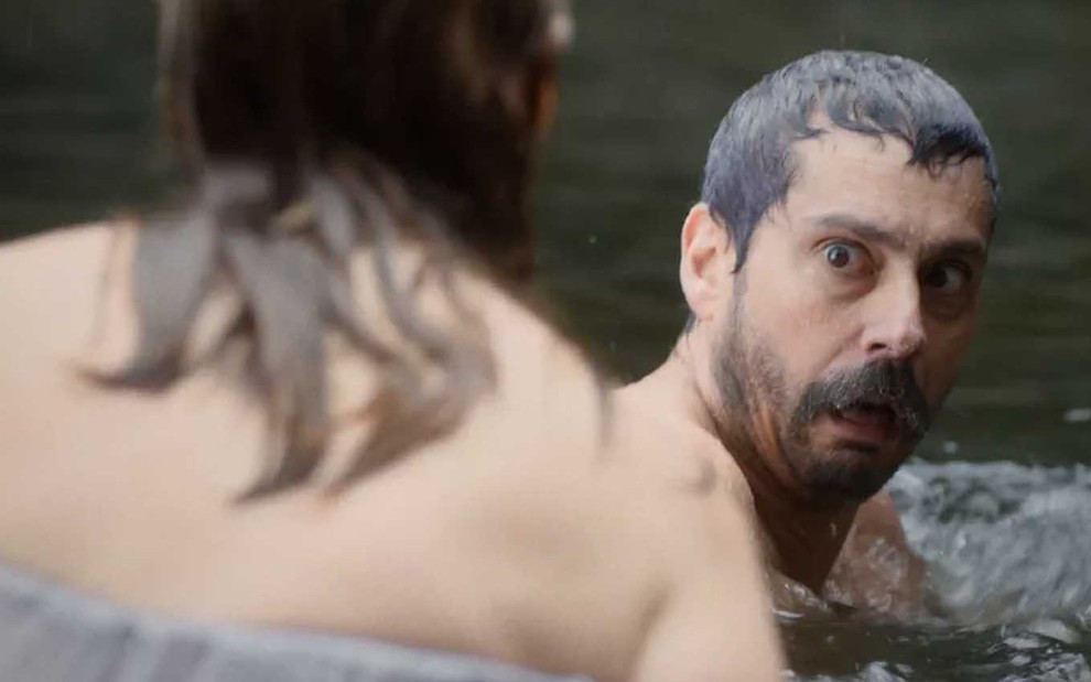 O ator Alexandre Nero está nu, dentro de um rio, e olha espantado para uma mulher em trajes sumários que está de costas do lado esquerdo em cena de Nos Tempos do Imperador