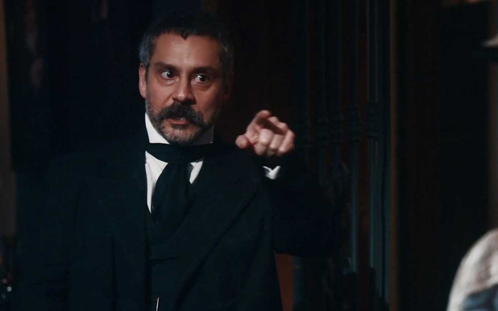 O ator Alexandre Nero caracterizado como Tonico com expressão de raiva enquanto aponta o dedo indicador da mão direita para a direita em cena de Nos Tempos do Imperador