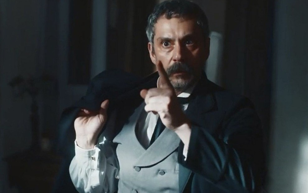 O ator Alexandre Nero com o dedo indicador da mão direita em riste como o Tonico em cena de Nos Tempos do Imperador