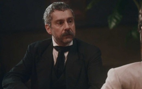 O ator Alexandre Nero caracterizado com Tonico sentado à mesa e com expressão de poucos amigos em cena de Nos Tempos do Imperador