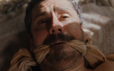 O ator Alexandre Nero, em um close-up no rosto, amordaçado com um pedaço de pano como o Tonico em cena de Nos Tempos do Imperador
