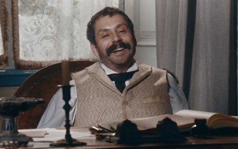 O ator Alexandre Nero com expressão de prazer como Tonico sentado à mesa de um escritório em cena de Nos Tempos do Imperador