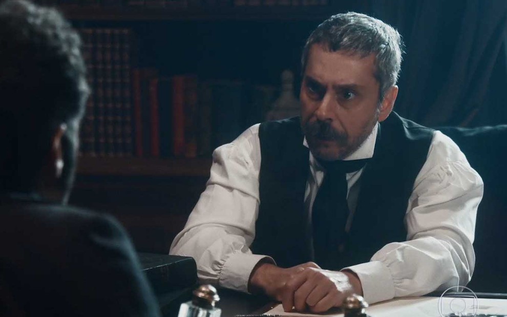 O ator Alexandre Nero sentado à mesa, com expressão de raiva, como o Tonico em cena noturna de Nos Tempos do Imperador