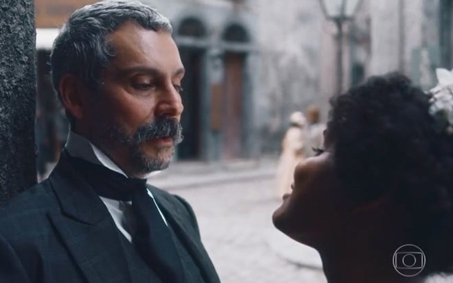 Tonico Rocha (Alexandre Nero) está encostado em parede e conversa com Zayla (Heslaine Vieira) em cena de Nos Tempos do Imperador