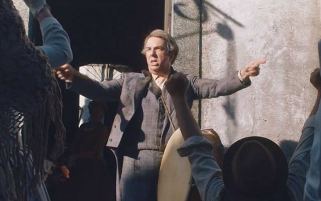 O ator Augusto Madeira como Quinzinho no meio de uma praça, com um bumbo e de braços abertos em cena de Nos Tempos do Imperador