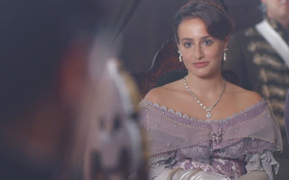 Leopoldina (Bruna Griphao) está sentada em banco da igreja entre pessoas, ela usa um vestido lilás em cena de Nos Tempos do Imperador