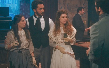 Leopoldina (Bruna Griphao), Pierre (Gabriel Falcão) e Isabel (Giulia Gayoso) estão em pé no cassino; as princesas seguram taças em cena de Nos Tempos do Imperador