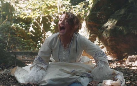 A atriz Bruna Griphao está caída no meio de uma clareira com um vestido branco como Leopoldina e grita por socorro como a Leopoldina em cena de Nos Tempos do Imperador