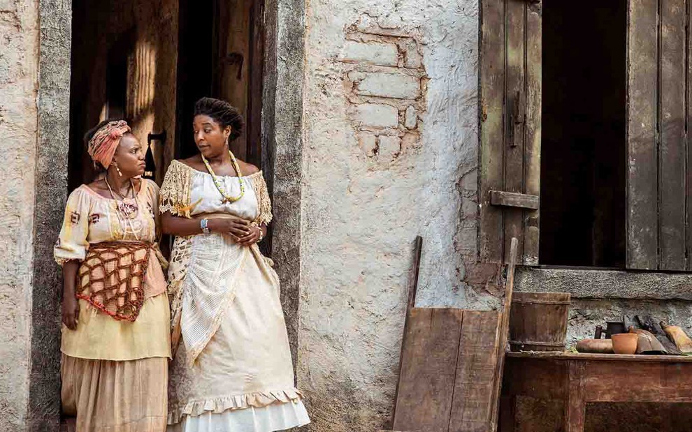 As atrizes Daniel Ornellas como Cândida e Mary Sheyla como Abena, com roupas com motivos africanos, típicas das classes populares do século 19, conversam na porta de uma casa humilde em cena de Nos Tempos do Imperador